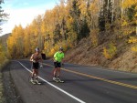 Two skiers rollerskiing up a winding mountain road lined with yellow aspens
