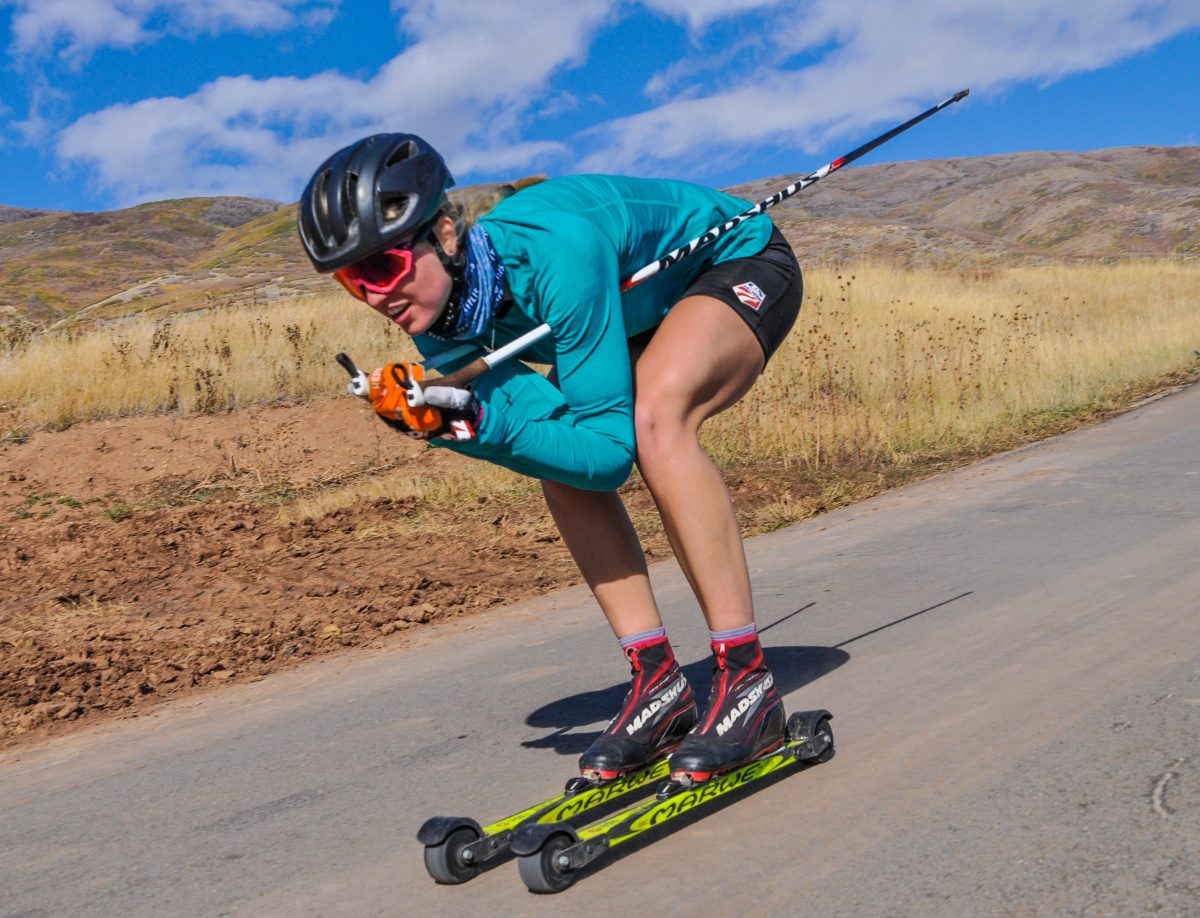 Hailey Swirbul in a teal top, black helmet and orange goggles crouched low on rollerskis on a paved road with dry grassy hills behind