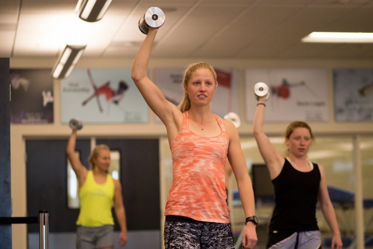 Three athletes pressing dumbbells overhead during a gym strength session