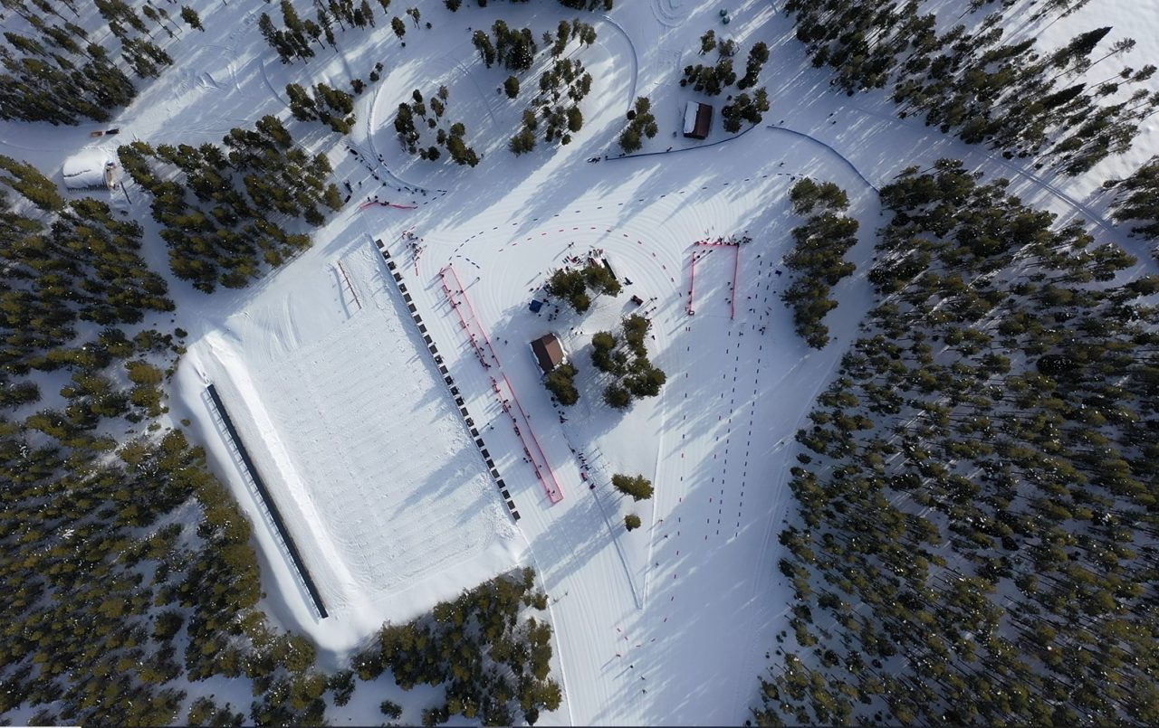 Aerial view of a snow-covered ski stadium and biathlon range cut into a pine forest, with groomed trails leading away