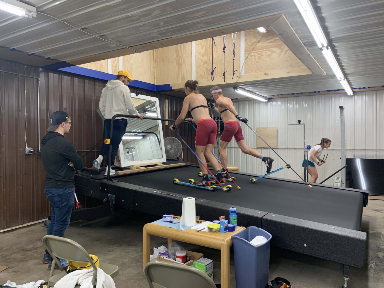 Two skiers in red shorts and heart rate straps skating on a wide rollerski treadmill while two coaches watch from the side