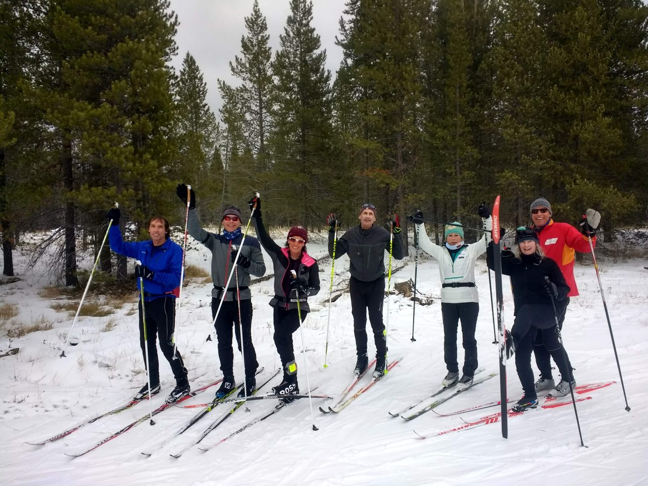 A group of eight cross-country skiers raising their poles and smiling on a snowy trail among pine trees