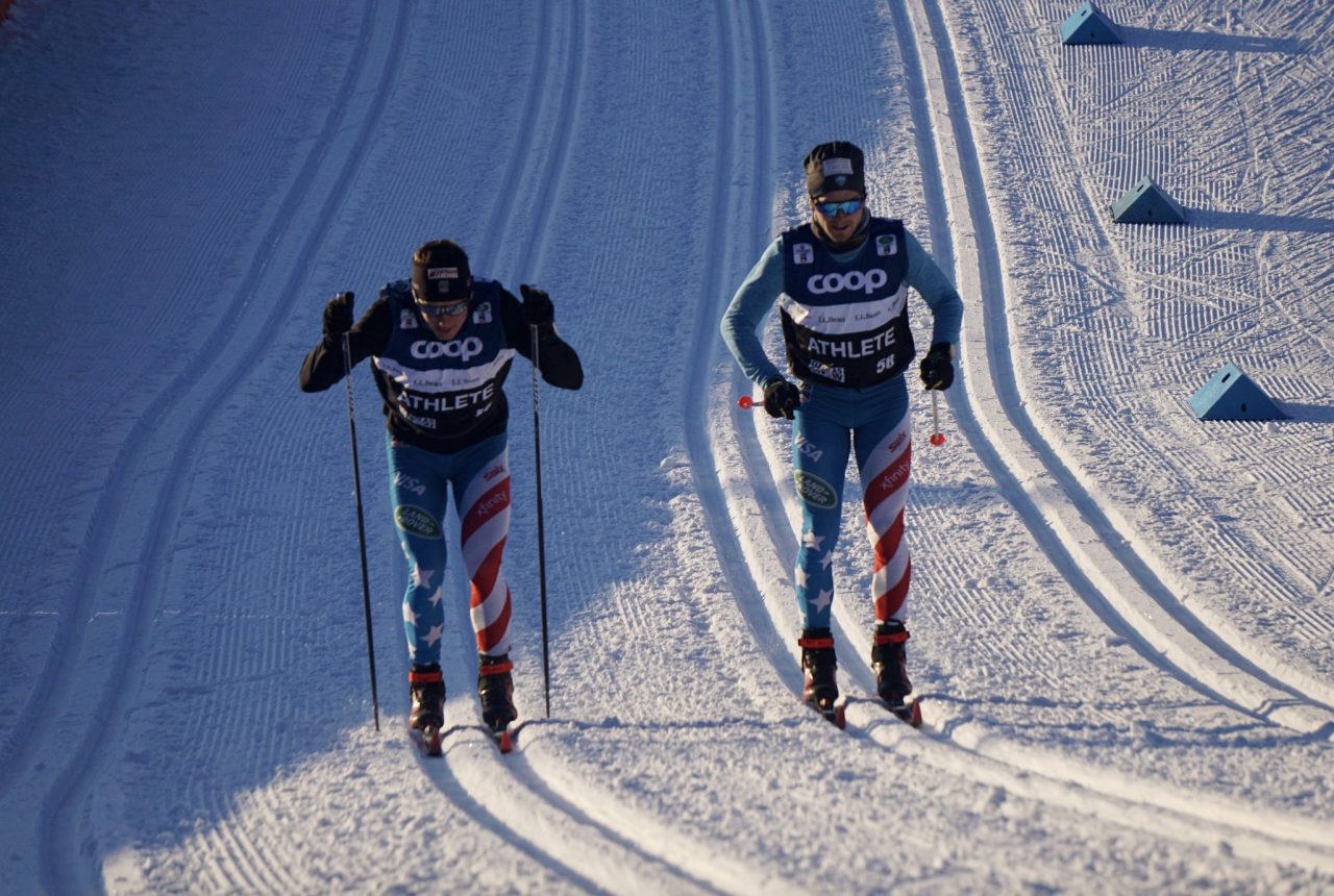 Two skiers in U.S. suits and Coop athlete bibs striding side by side up groomed classic tracks in low winter sun