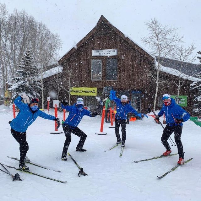 Four coaches on skis in falling snow outside a nordic center lodge