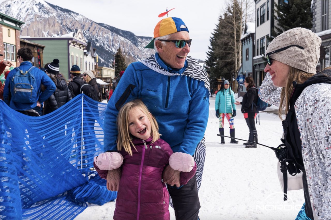 An older skier in a propeller cap laughs with a young girl on a snow-covered town street