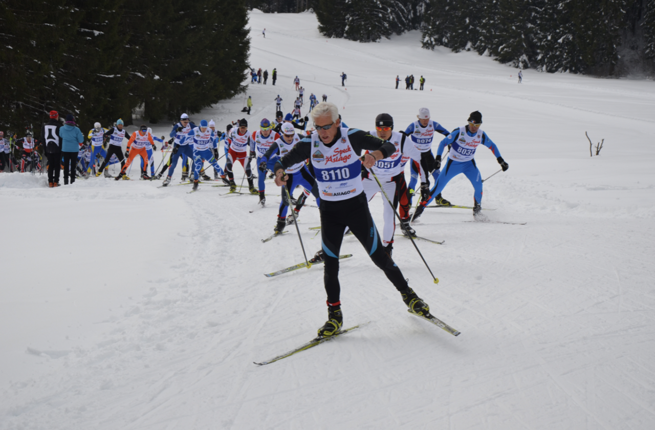 A masters field skating up a snowy climb behind a white-haired leader wearing bib 8110