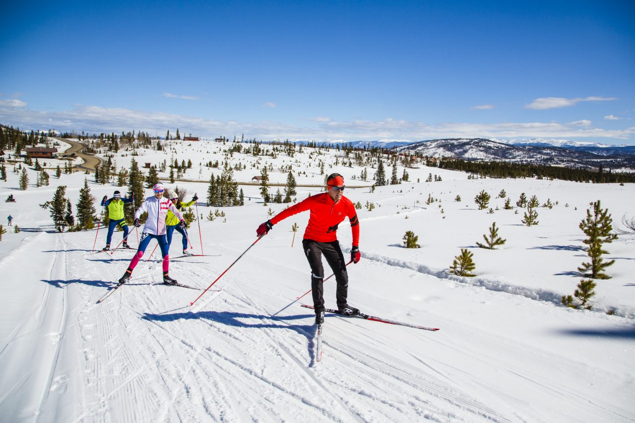 A skate skier in a red jacket leads three others along a groomed trail across open, snow-covered rolling terrain.