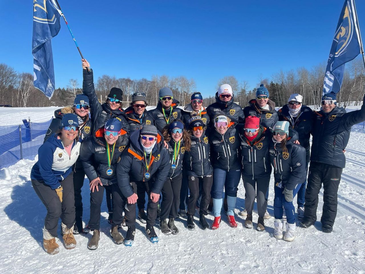 The St. Scholastica ski team in navy Saints jackets and sunglasses posing on snow under blue sky, several holding NCAA medals, with two blue team flags raised behind them.