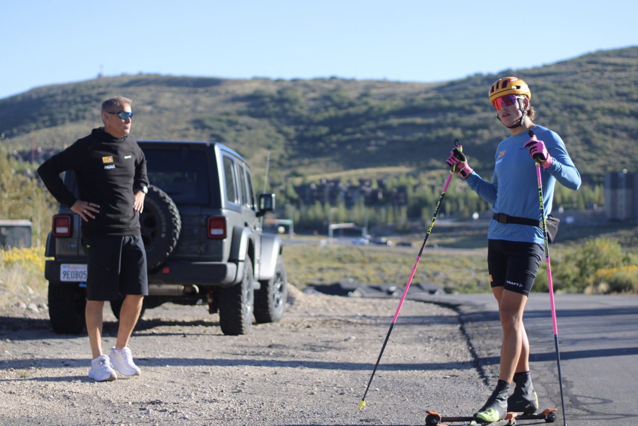 A skier in a blue long-sleeve top, yellow helmet and pink poles stands on rollerskis beside a man in black next to a gray Jeep, with sage-covered hills behind