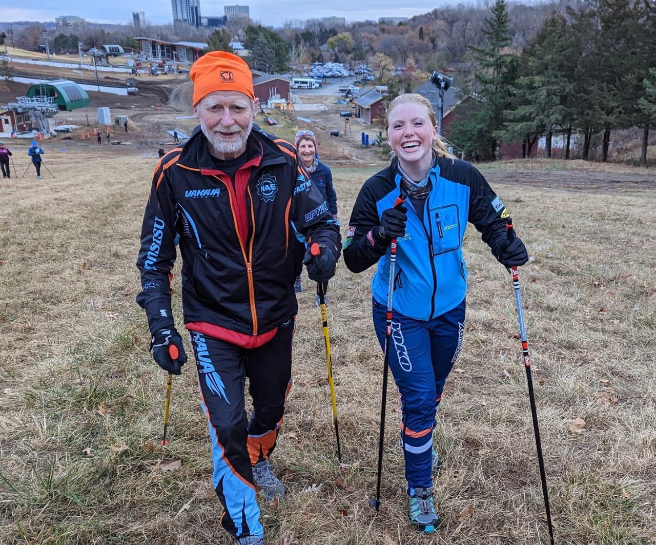 An older skier in a Vakava race suit walks up a grassy hill with poles beside a younger skier