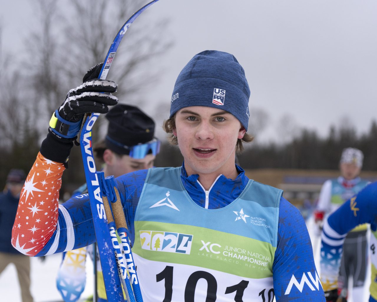 Junior skier Logan Drevlow in a Midwest race suit and bib 101 holding his skis at Junior Nationals