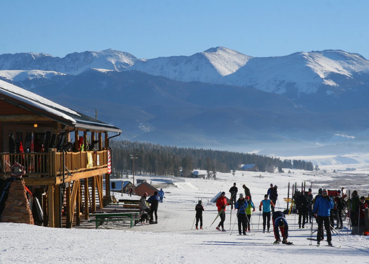 Cross-country skiers gathered on the snow outside a wooden Nordic center lodge, with snow-covered peaks rising behind.