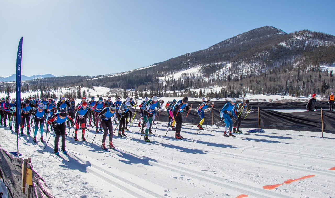 A long line of cross-country skiers lined up across a groomed trail at the start of a mass start race below a forested hill.