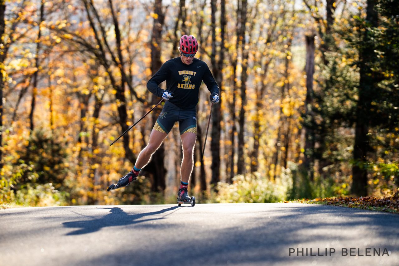 A University of Vermont skier rollerskiing up a paved climb through autumn woods
