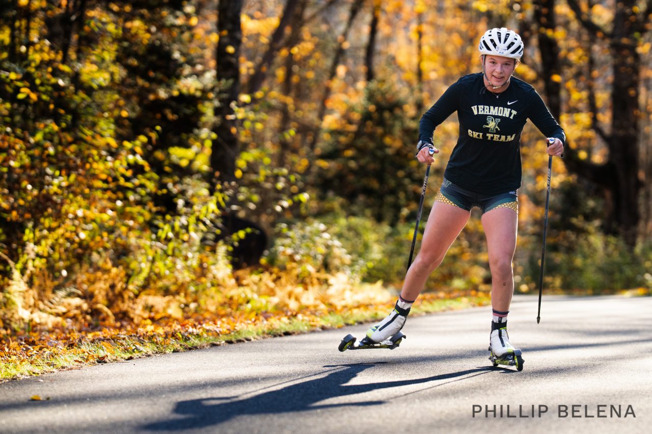 A skier in a Vermont jacket and white helmet skating on rollerskis along a paved path lined with autumn trees