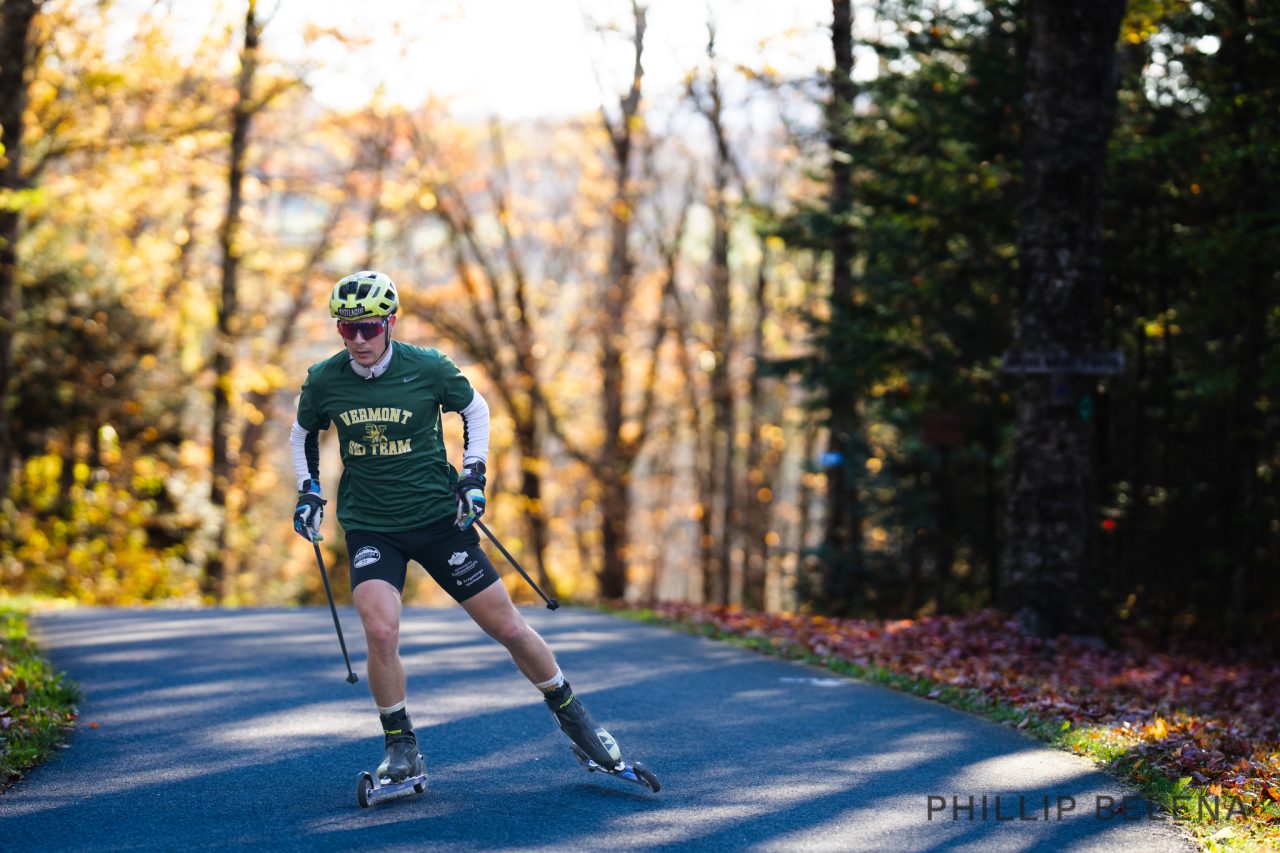 A skier in a green Vermont shirt and helmet skating on rollerskis down a paved path through autumn woods