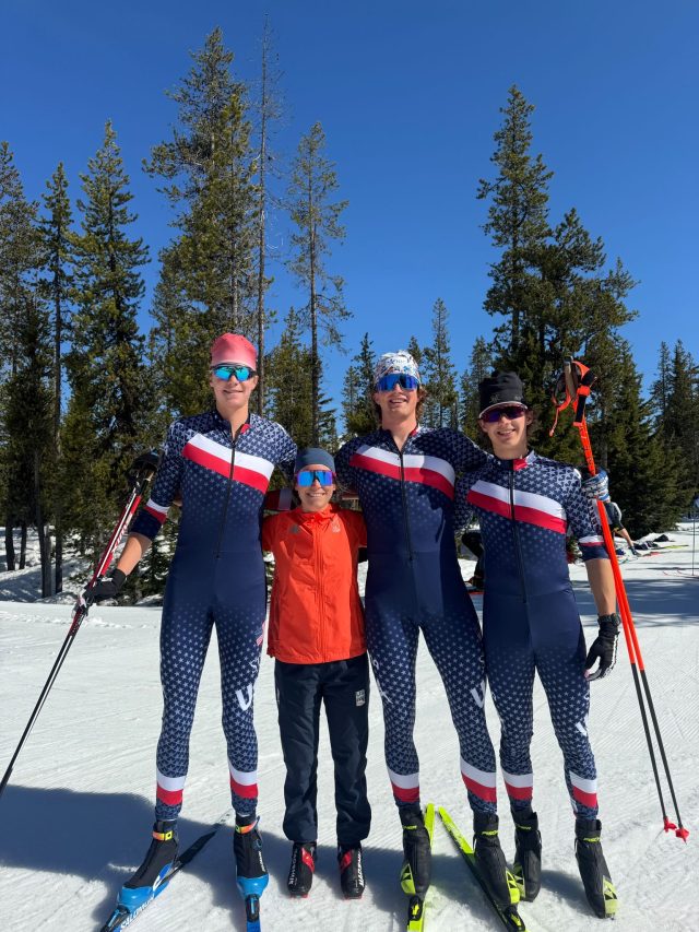 Three skiers in U.S. team suits and one in an orange jacket standing together on snow among pine trees