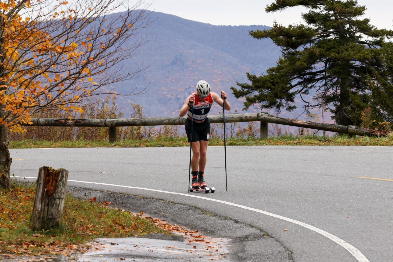A rollerskier in a red and black suit climbing a paved mountain road past autumn trees
