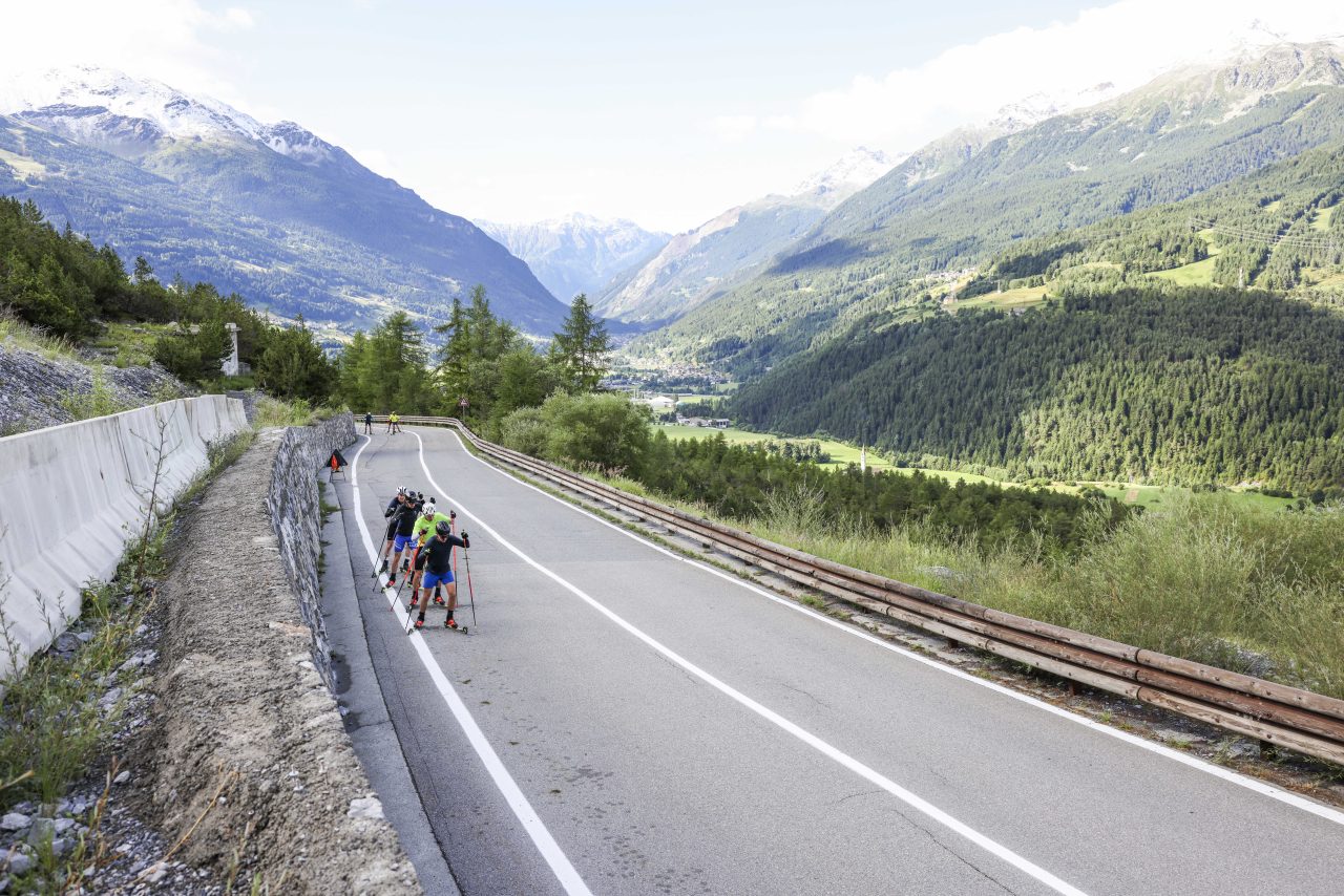 Four skiers rollerskiing in a line up a switchback road climbing out of a green alpine valley
