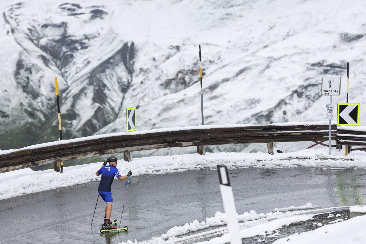 A rollerskier in a blue top climbs a wet mountain road edged with fresh snow beneath steep snowy slopes