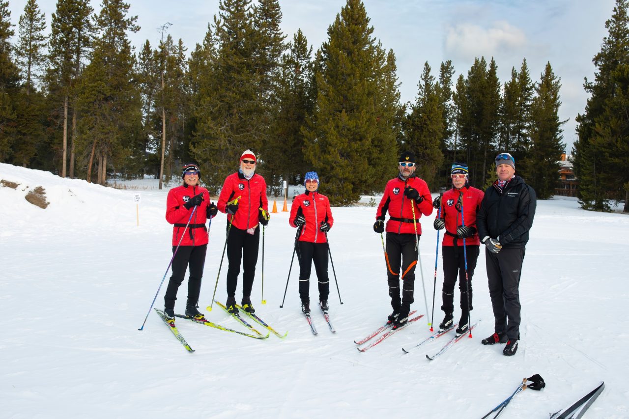 Six skiers in matching red jackets and one man in a black jacket pose together on skis in a snowy clearing