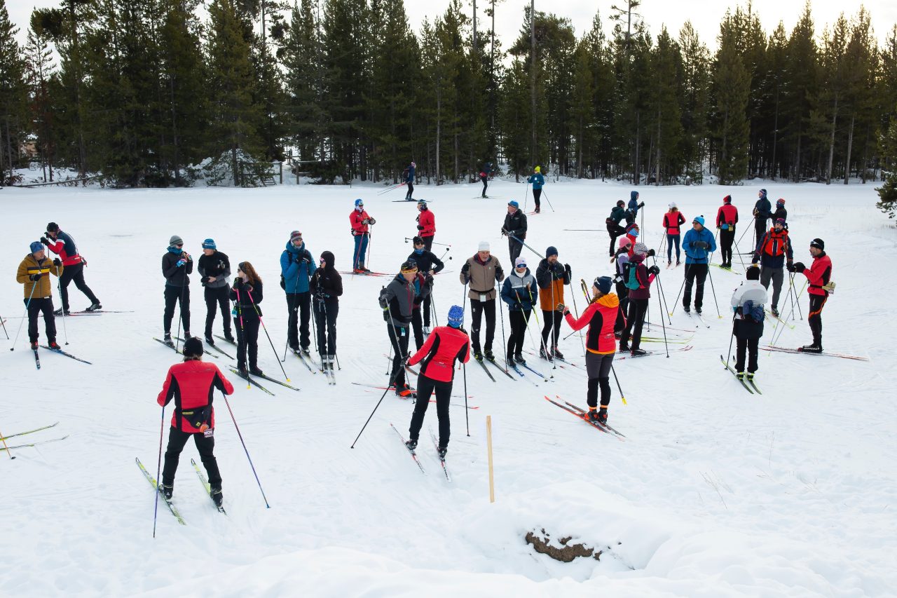 A large group of cross-country skiers and coaches in red jackets gathered in clusters on a snowy field ringed by pine trees