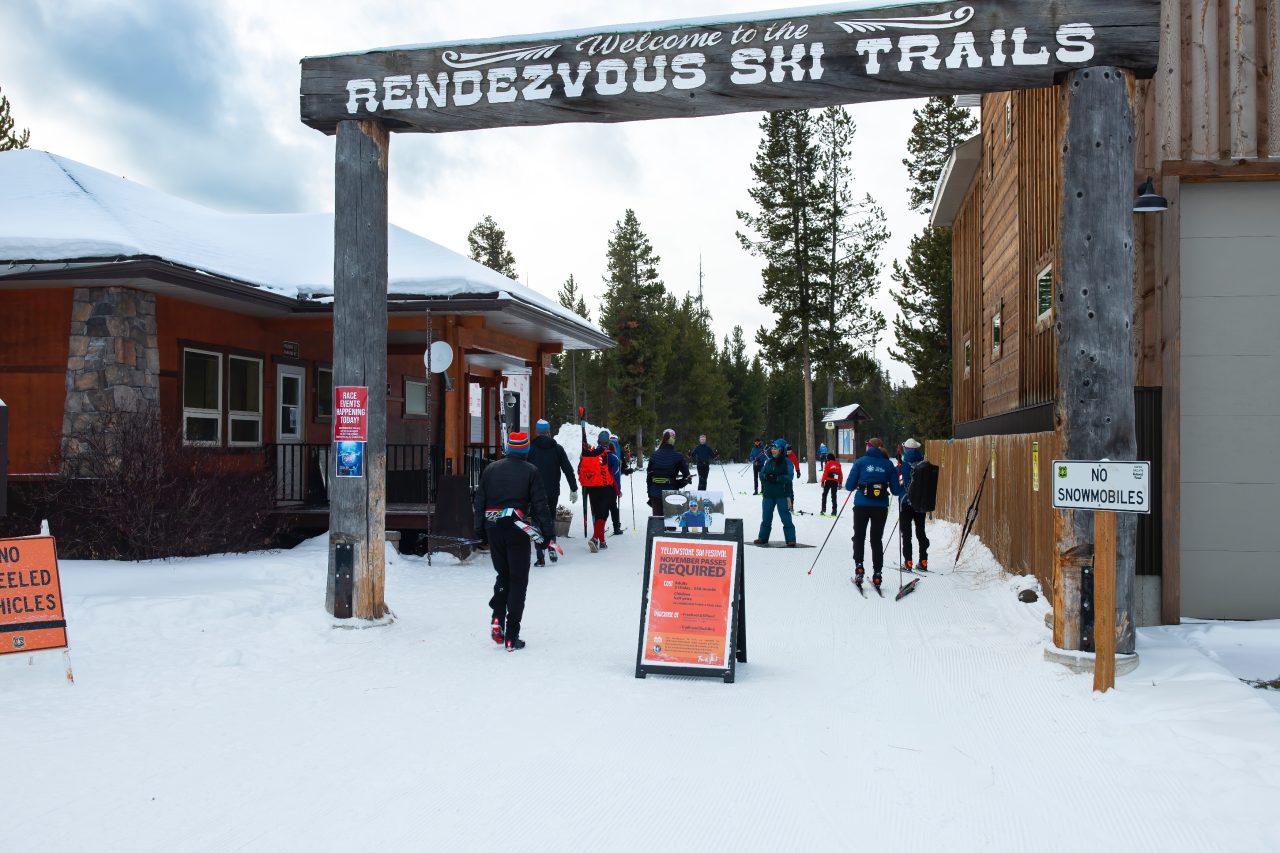 Skiers walking and skiing under a carved wooden arch reading Welcome to the Rendezvous Ski Trails, with a sign saying November passes required