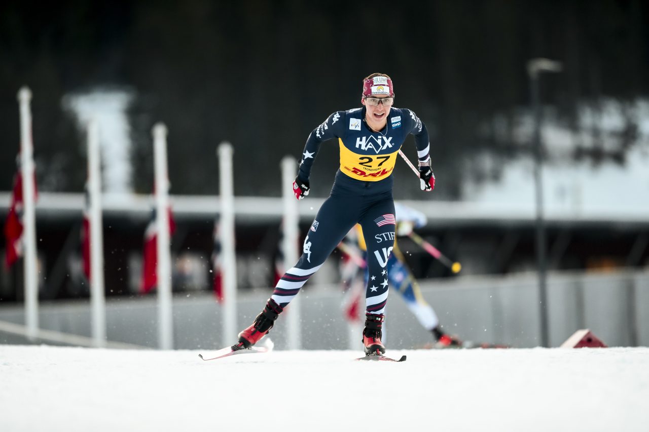 Julia Kern in a U.S. suit and yellow bib skating toward the camera past a row of flags