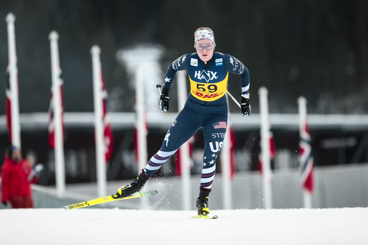 Kate Oldham in a U.S. team suit and yellow bib 59 skating toward the camera past a row of flags
