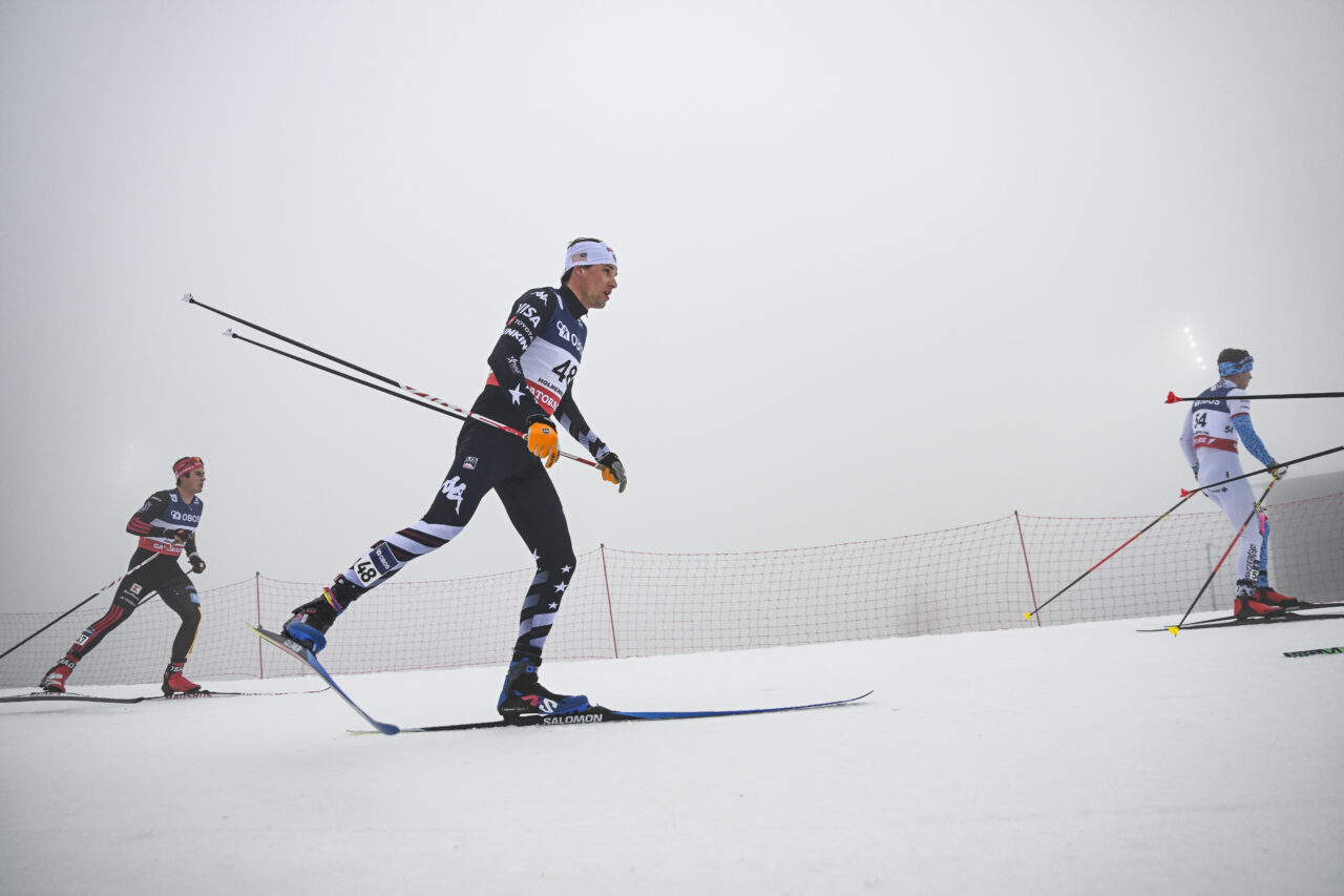 Kevin Bolger in a U.S. suit skating downhill in fog with other racers and trees behind