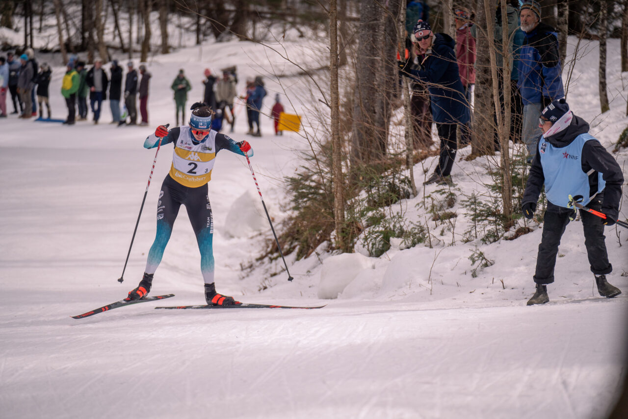 Luci Anderson in bib 2 classic skiing past spectators in the woods at Craftsbury