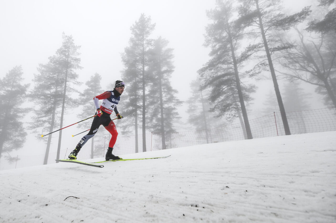 Thomas Stephen in a red and black Canada suit skating up a snowy slope with pines fading into fog