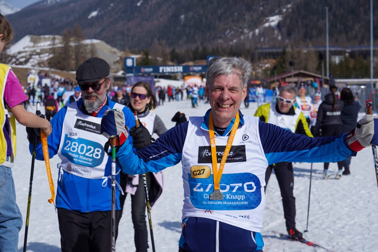 A grey-haired skier spreads his arms in celebration wearing a finisher medal at the Engadin Skimarathon finish