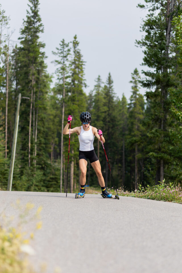 Alison Mackie rollerskiing toward the camera on a paved road lined with evergreens.