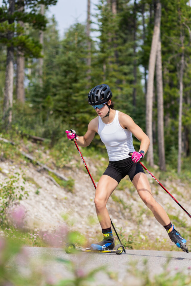 Alison Mackie skate rollerskiing uphill on a paved forest road in a white training top and black helmet.