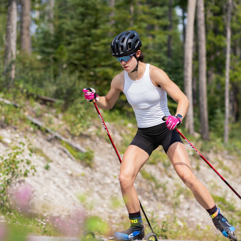Alison Mackie skate rollerskiing on a forest road in a white training top and black helmet.