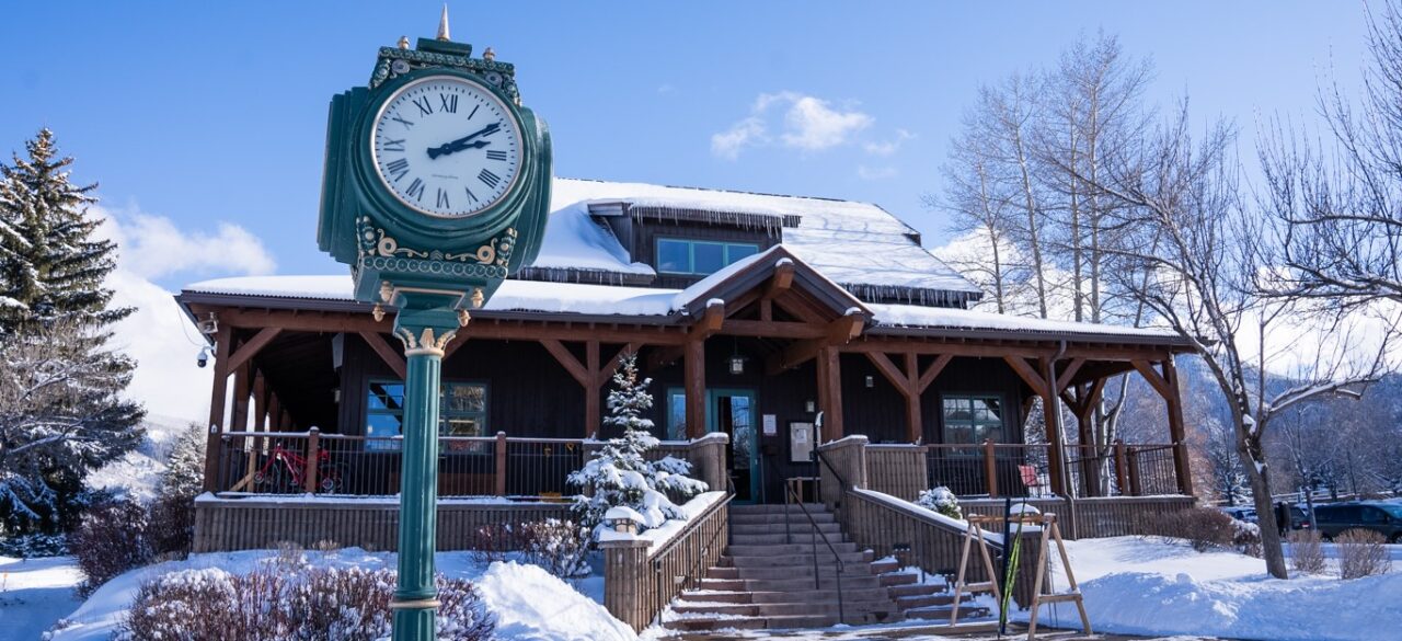A timber lodge with a snow-covered roof behind a green cast-iron clock tower, with steps leading up to the entrance on a clear winter day.