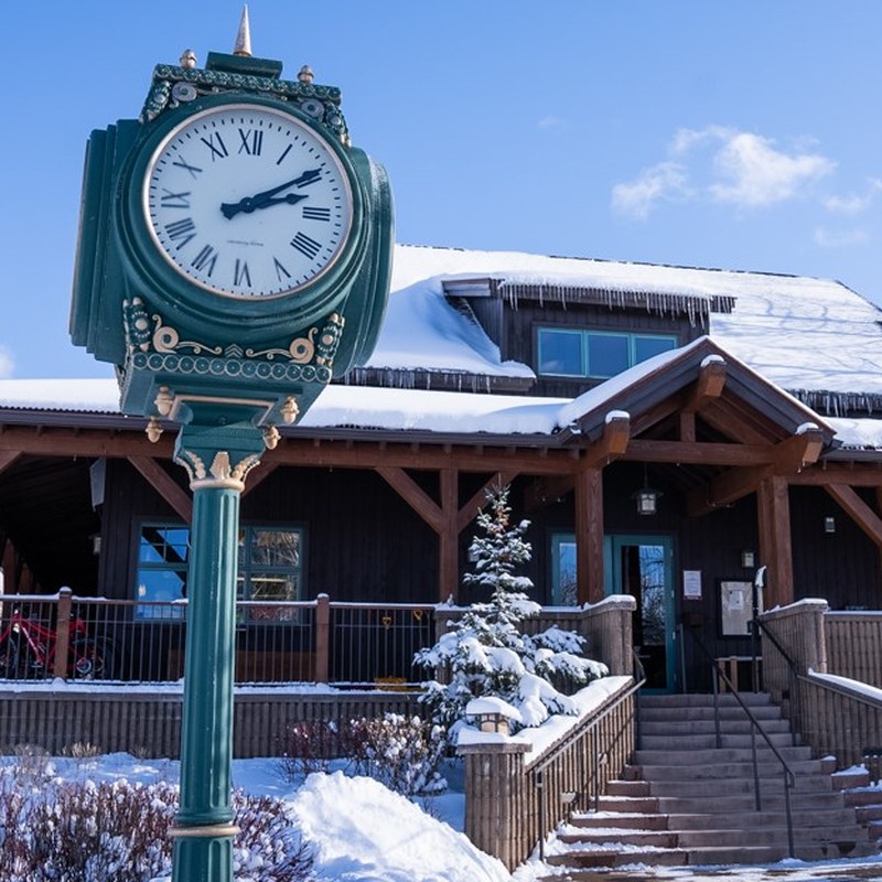 A green cast-iron clock tower standing in the snow in front of the timber lodge of the Aspen Cross Country Center.