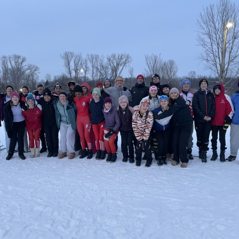 Members of the Benilde-St. Margaret's/DeLaSalle Nordic team stand together on a snowy field at dusk, several in red race suits.