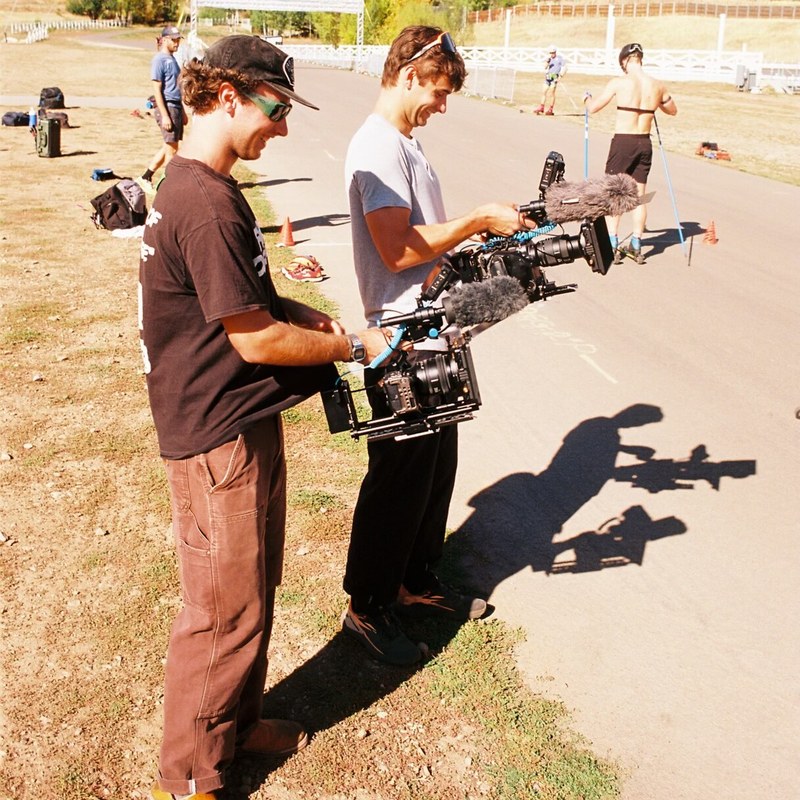 Two filmmakers stand in profile at the edge of a paved roller ski loop, each holding a cinema camera with a microphone mounted on top.