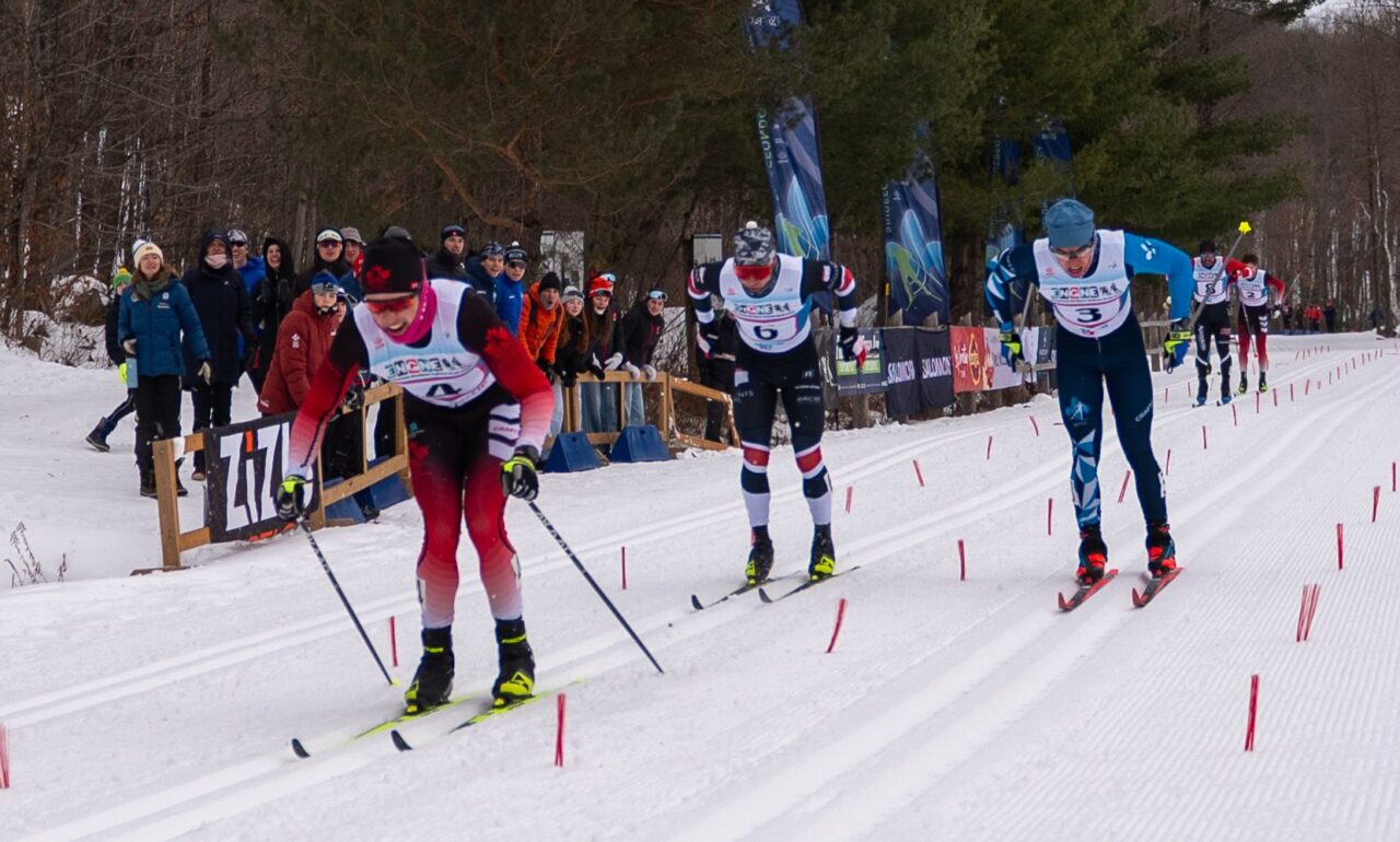 Three cross-country skiers double-poling side by side down a finishing straight, with spectators watching from behind a fence.
