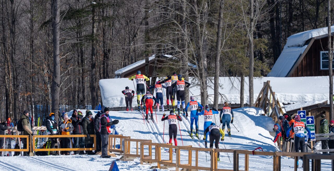 A line of racers climbing a hill past a wooden chalet while spectators watch from behind a fence.