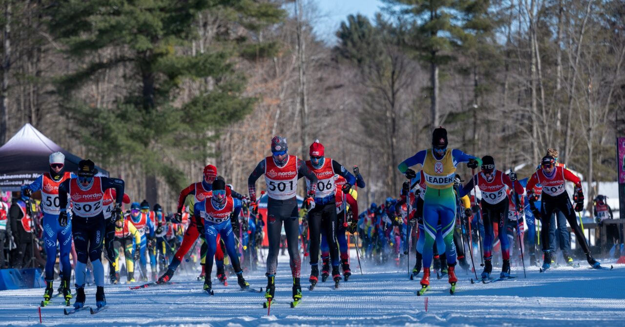 A wide field of skate skiers spread across a groomed start straight at the beginning of a mass start race.