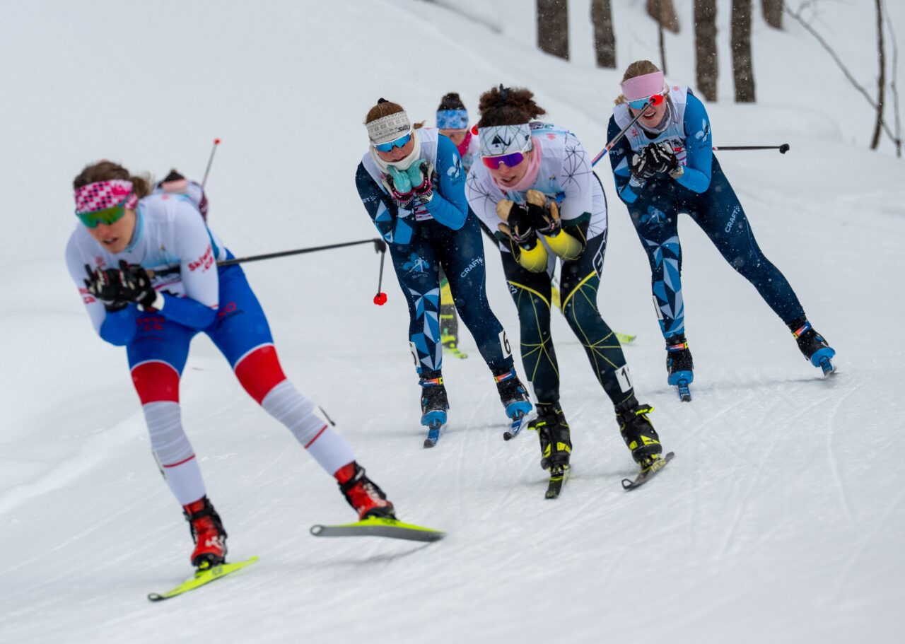 Five women skiers in a tight group tucking down a hill while snow falls around them.