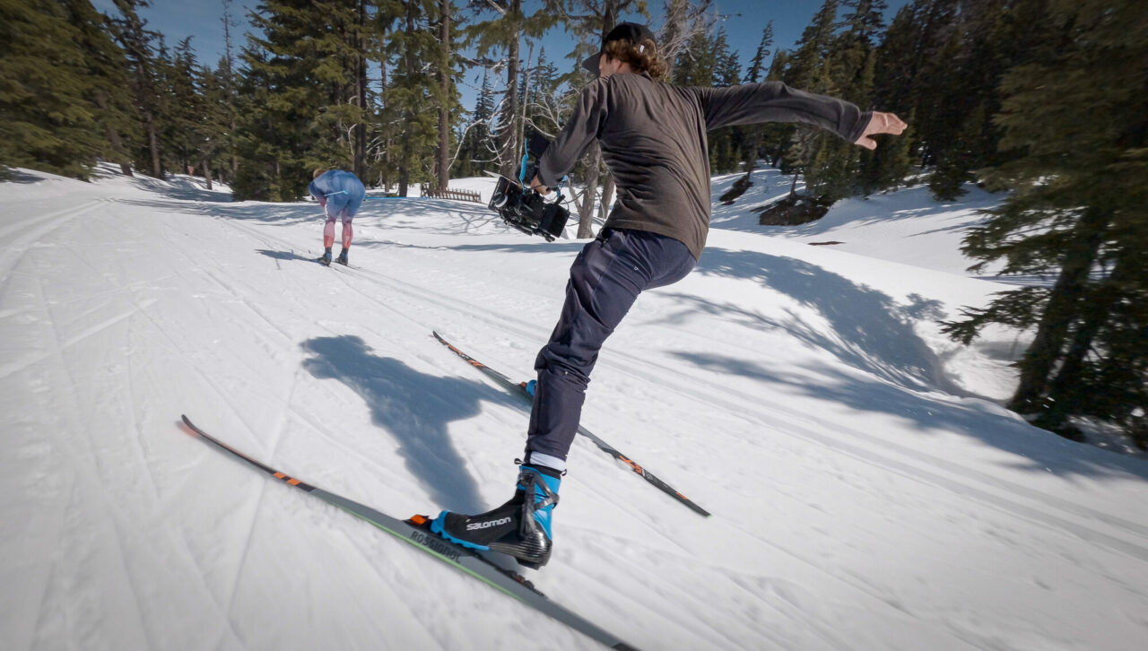 A filmmaker mid-stride on cross country skis carrying a cinema camera rig, with an athlete skiing ahead of him on a groomed snow trail.