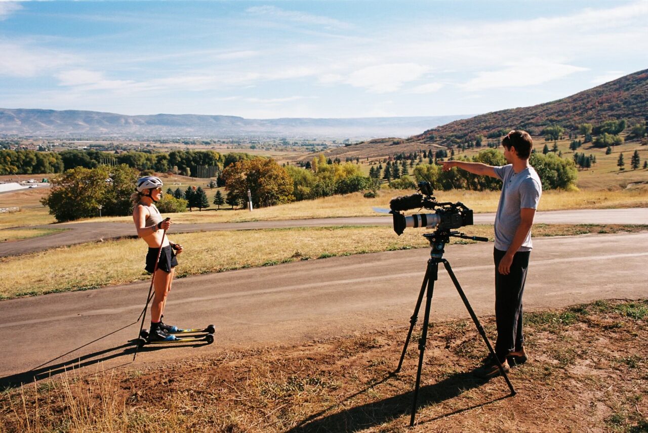 A skier on roller skis stands on a paved path while a filmmaker directs from behind a tripod-mounted cinema camera, with a wide valley behind them.