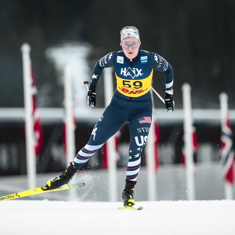 Kate Oldham in a U.S. team suit and yellow bib 59 skating toward the camera past a row of flags