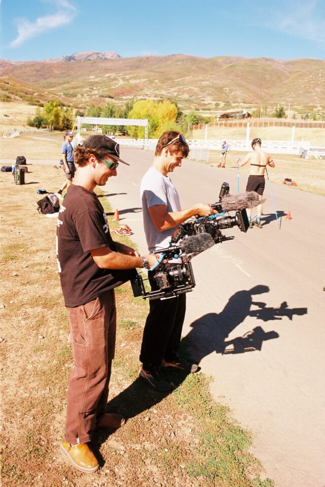 Two filmmakers stand in profile at the edge of a paved roller ski loop, each holding a cinema camera with a microphone mounted on top.