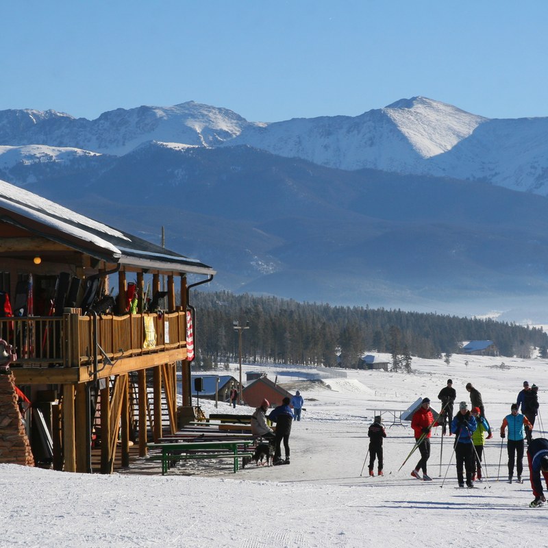 Cross-country skiers gathered on the snow beside a wooden Nordic center lodge, with snow-covered peaks behind.