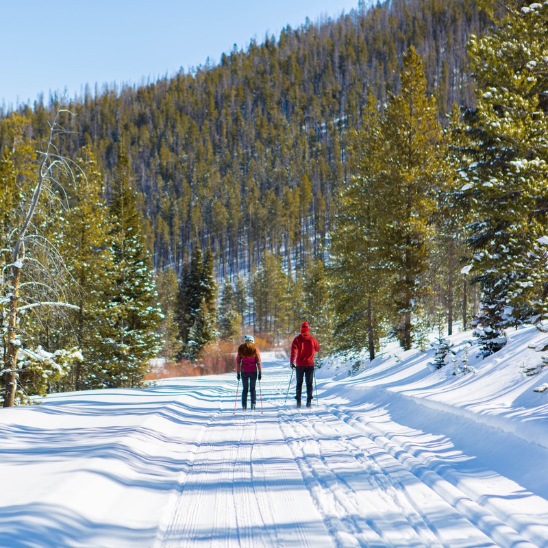 Two skiers on a groomed trail between snowy pines at Snow Mountain Ranch, one in a red jacket and one in a pink jacket.
